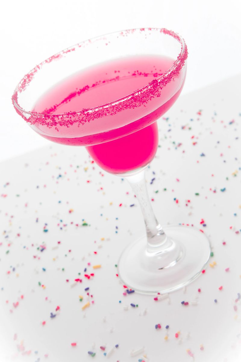 A hot pink margarita in a margarita glass with pink sugar on the rim; festive sprinkles surround it
