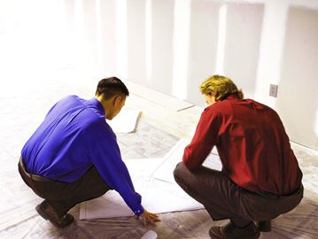Two men in colorful shirts reviewing blueprints on the floor.
