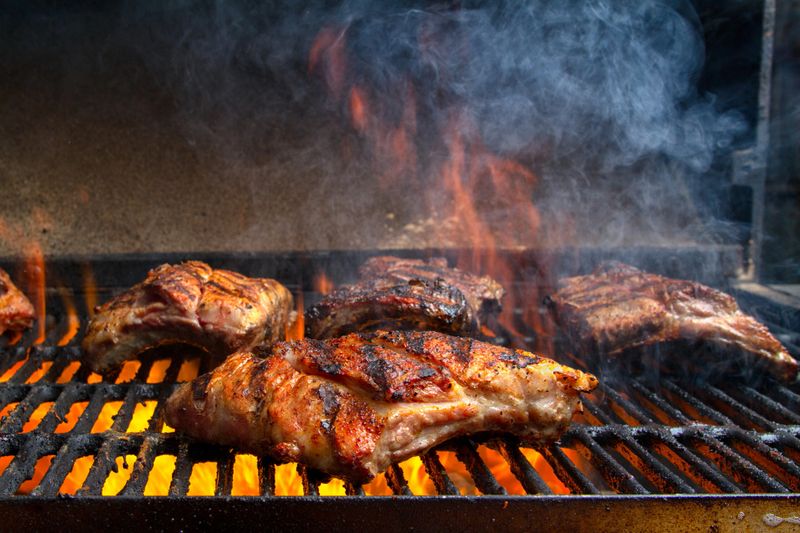 Grouping of Baby Back Ribs on a barbecue grill with flames leaping up to sear the meat sealing in the juices.  Ribs have nice grill marks and are seasoned with a dry rub and are almost ready to serve.