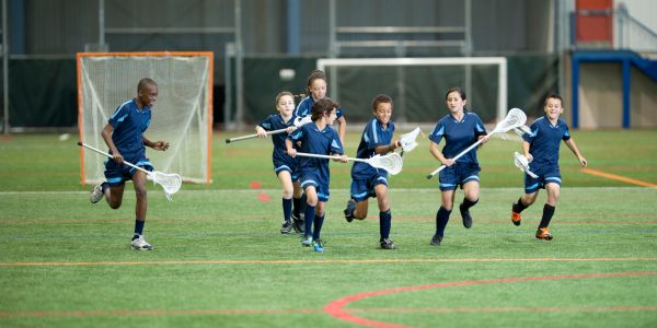 Children in blue uniforms running on a lacrosse field with sticks.