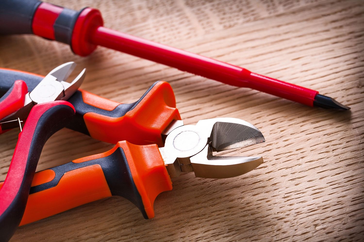 Close-up of orange and black pliers with a red screwdriver on a wooden surface.