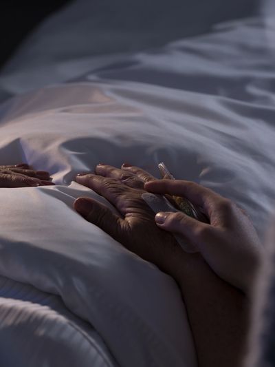 A hand holding an elderly hand on a hospital bed with an IV line.