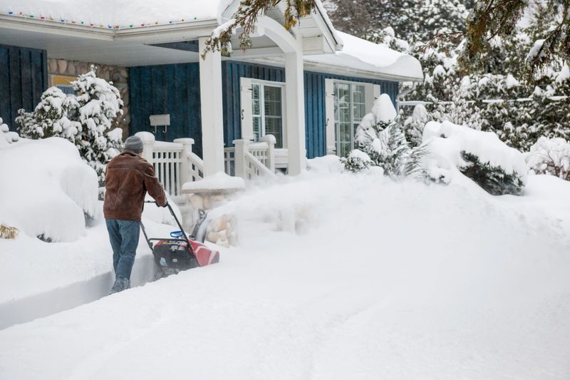 Man using snowblower to clear deep snow on driveway near residential house after heavy snowfall