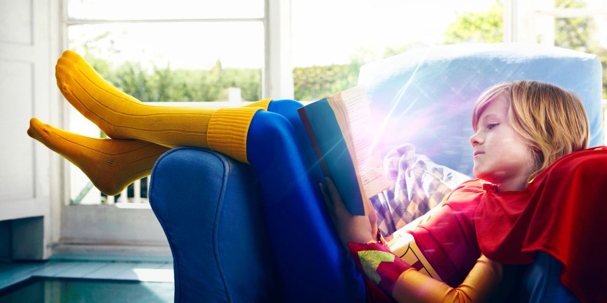 Child in superhero costume reading a glowing book on a blue couch.