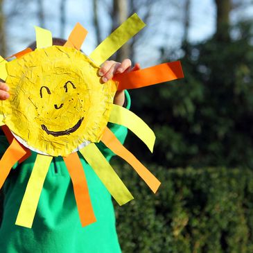 Child holding a handmade smiling sun craft with colorful rays outdoors.