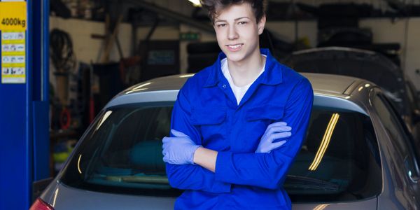 Young mechanic in blue uniform standing in front of a car in a garage.