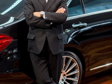 Man in black suit leaning against a luxury car.