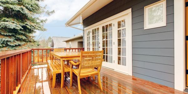 Wooden patio with table and chairs on a wet deck beside blue house.