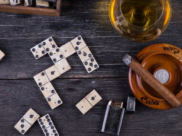 Dominoes, a Cuban cigar, lighter, and a glass of drink on a wooden surface.