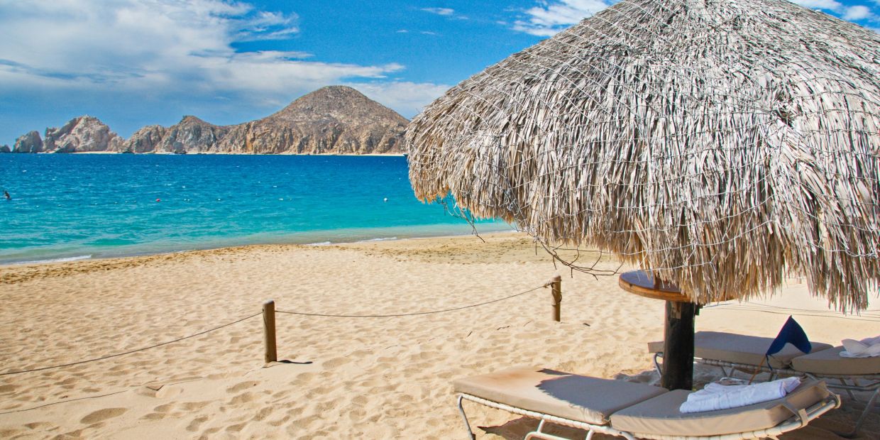 Beach scene with lounge chairs under a straw umbrella and ocean view.
