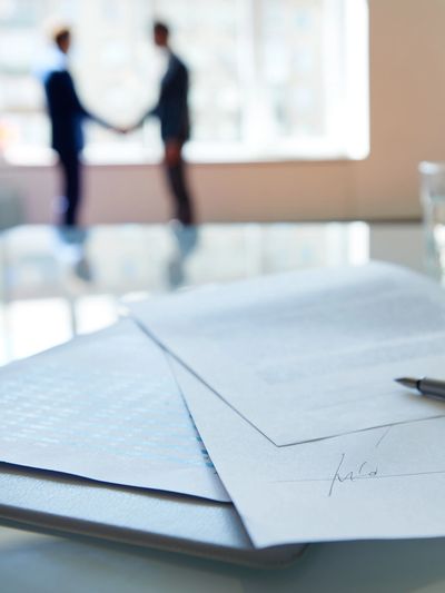 Signed documents and pen on desk with two people shaking hands blurred in background.