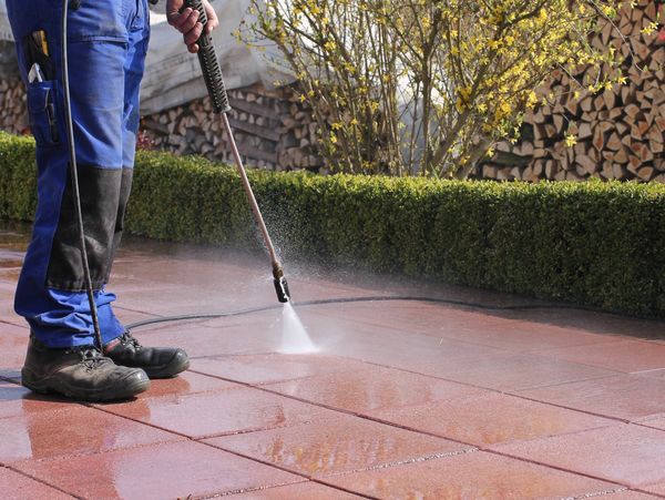 Person power washing outdoor tiles near a garden hedge.