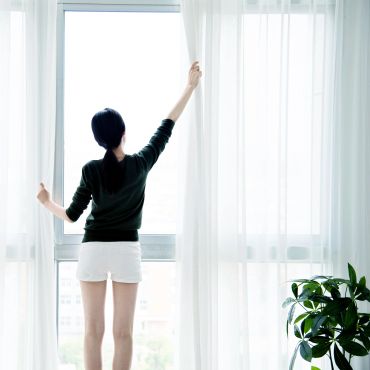 Person opening white curtains to bright daylight in a modern room.