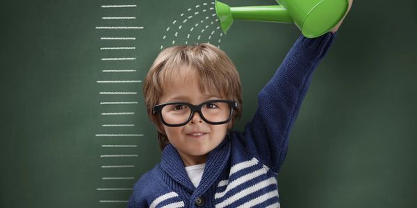 Child wearing glasses 'watering' his head with a green watering can, standing in front of a height chart.