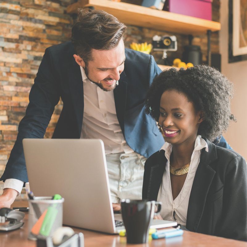 Caucasian male is helping his black female colleague while she is working on laptop computer in the office. On the desk are pen holder with markers cup of coffee and pens, and brick wall is in the background.
