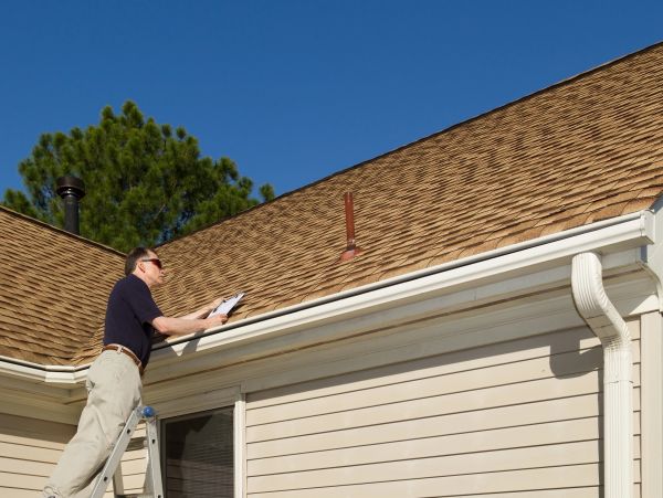 A man inspecting a house roof and gutters on a sunny day.