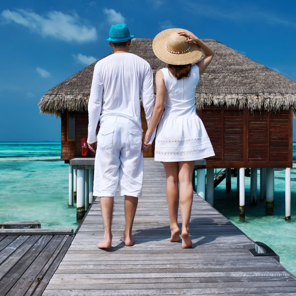 Couple holding hands on a wooden pier leading to a beach hut over turquoise water.