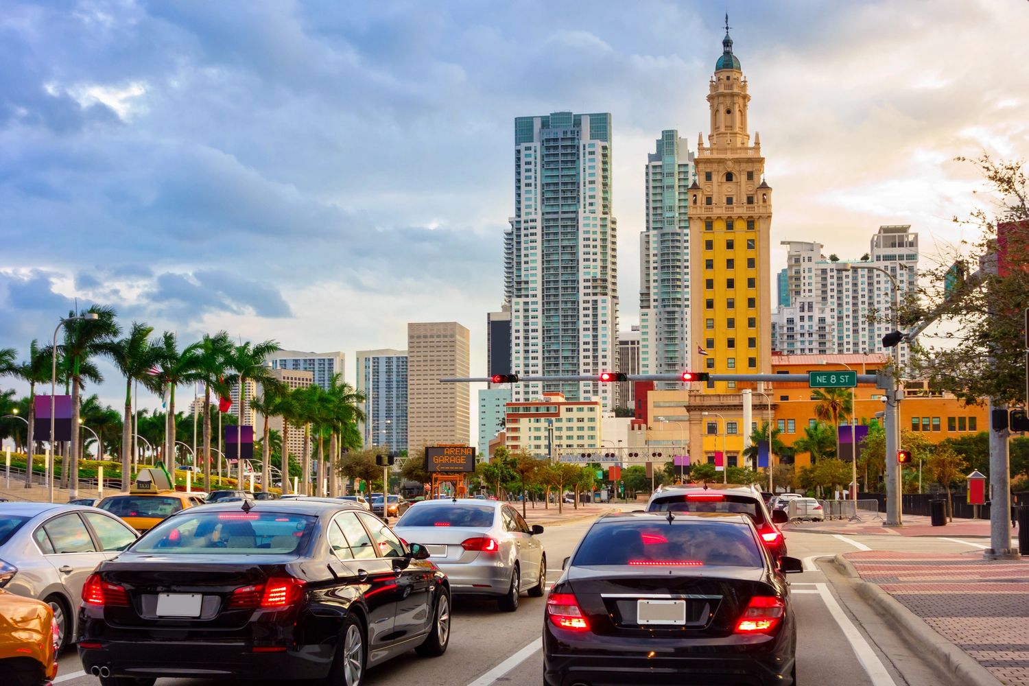 Busy city street with cars, palm trees, and tall buildings at sunset.