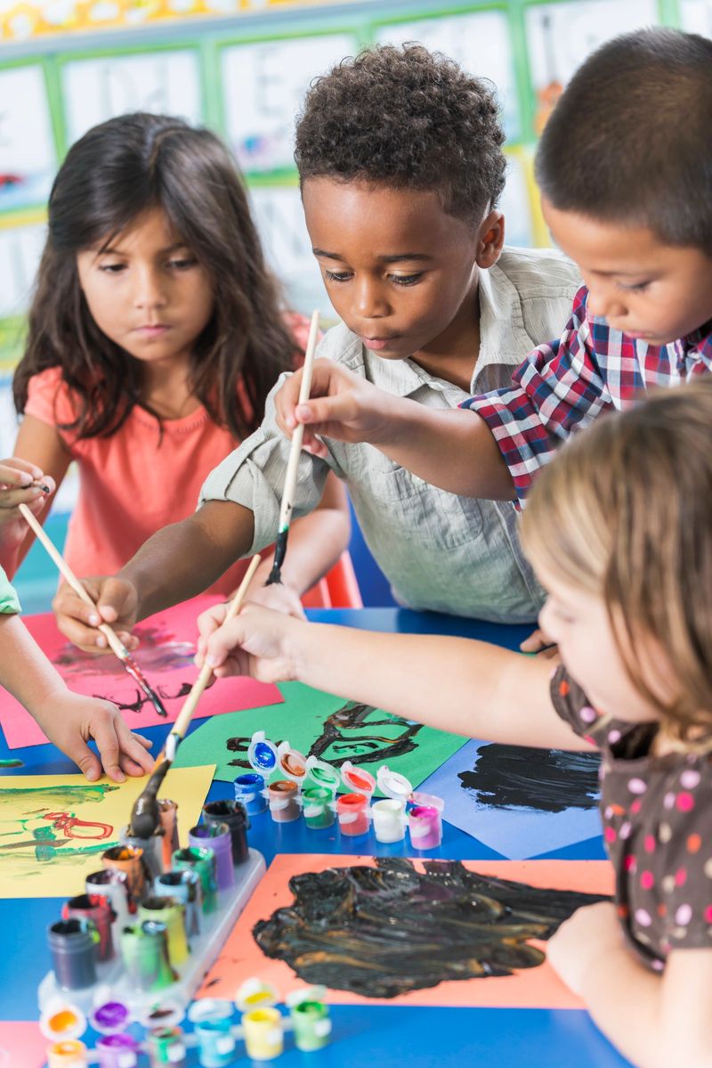 A group of preschool or kindergarten children in art class painting pictures at a table.  A little African American boy, 6 years old, is in the middle, reaching over to dip his brush in the paint.