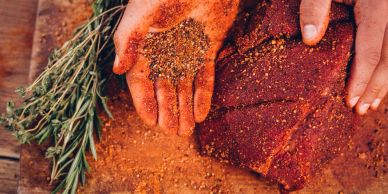 Hands seasoning a raw steak with herbs and spices.