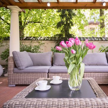 Outdoor patio with wicker sofa, pink tulips in a vase, and two coffee cups on a table.