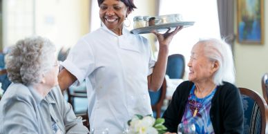 Caregiver serving two elderly women with a smile in a cozy setting.