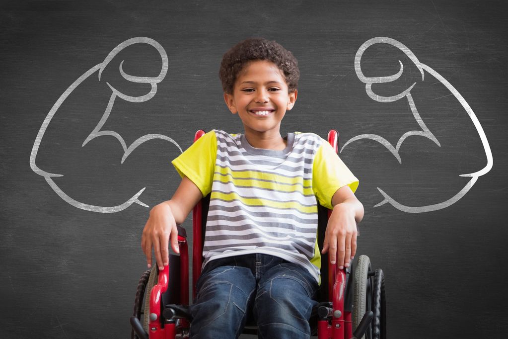 Smiling boy in wheelchair with chalk-drawn muscular arms on blackboard behind him.