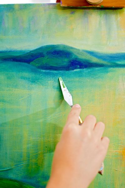 Hand using palette knife to paint a green and blue canvas.