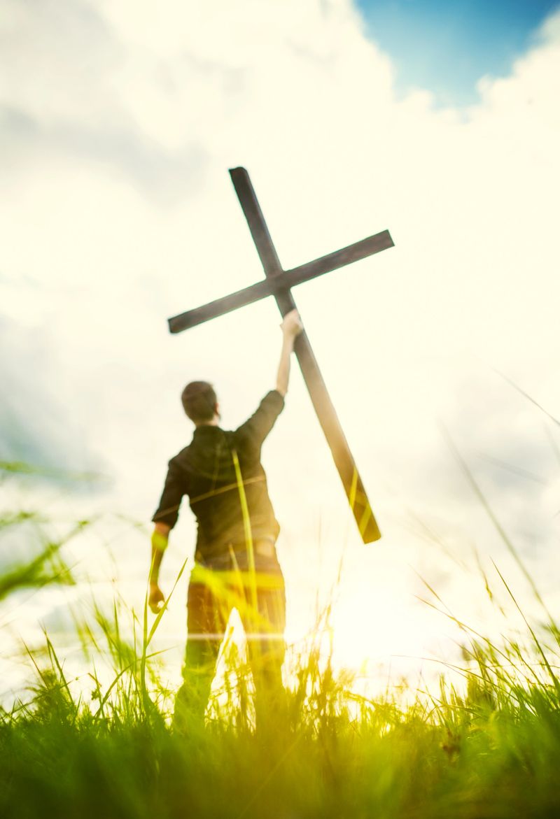 A man standing in a green grassy area surrounded by bright golden sunlight, lifting up a large wooden cross toward the Vertical image with copy space in the clouds.