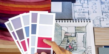 A hand holds paint swatches over architectural sketches and wood samples.