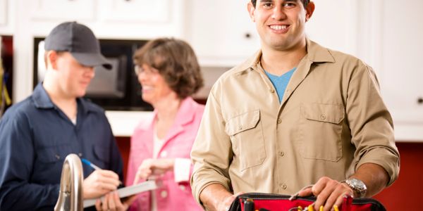 Smiling handyman with toolbox in foreground, two people discussing in background.
