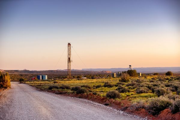 A man works near two trucks and a tall drilling rig in a grassy area under clear blue sky.