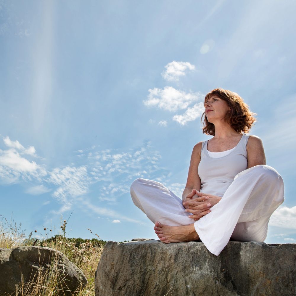 Woman meditating outdoors on a rock under a clear blue sky.