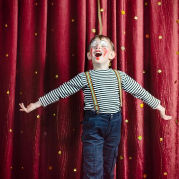 Child clown joyfully performing on stage with red curtain backdrop.