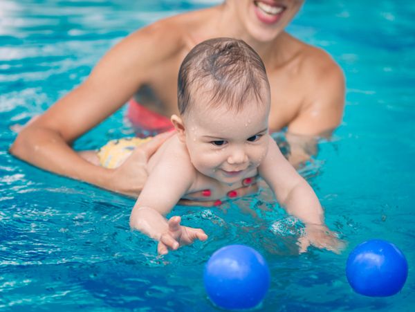 A baby enjoys swimming with an adult in a pool, reaching for blue balls.