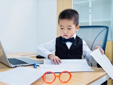 Young boy dressed formally working with papers at a desk with a laptop.