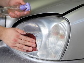 Person restoring a car's cloudy headlight with sandpaper and spray bottle.