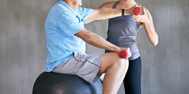 Elderly man exercising with dumbbells guided by a trainer on an exercise ball.