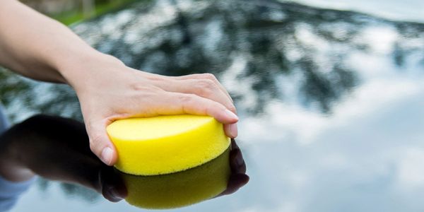 Hand using a yellow sponge to clean a shiny black car surface outdoors.