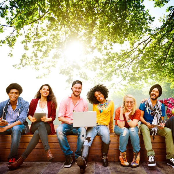 Group of diverse young adults sitting outdoors with digital devices, smiling under bright sunlight.