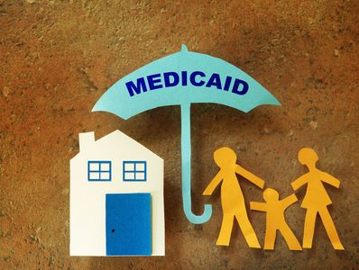 Paper cutouts show Medicaid protecting a family and home under an umbrella.