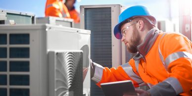 Technician inspecting HVAC unit on rooftop with tablet, wearing safety gear.