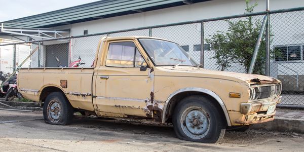 Old, rusty yellow pickup truck with flat tires parked beside a fence.