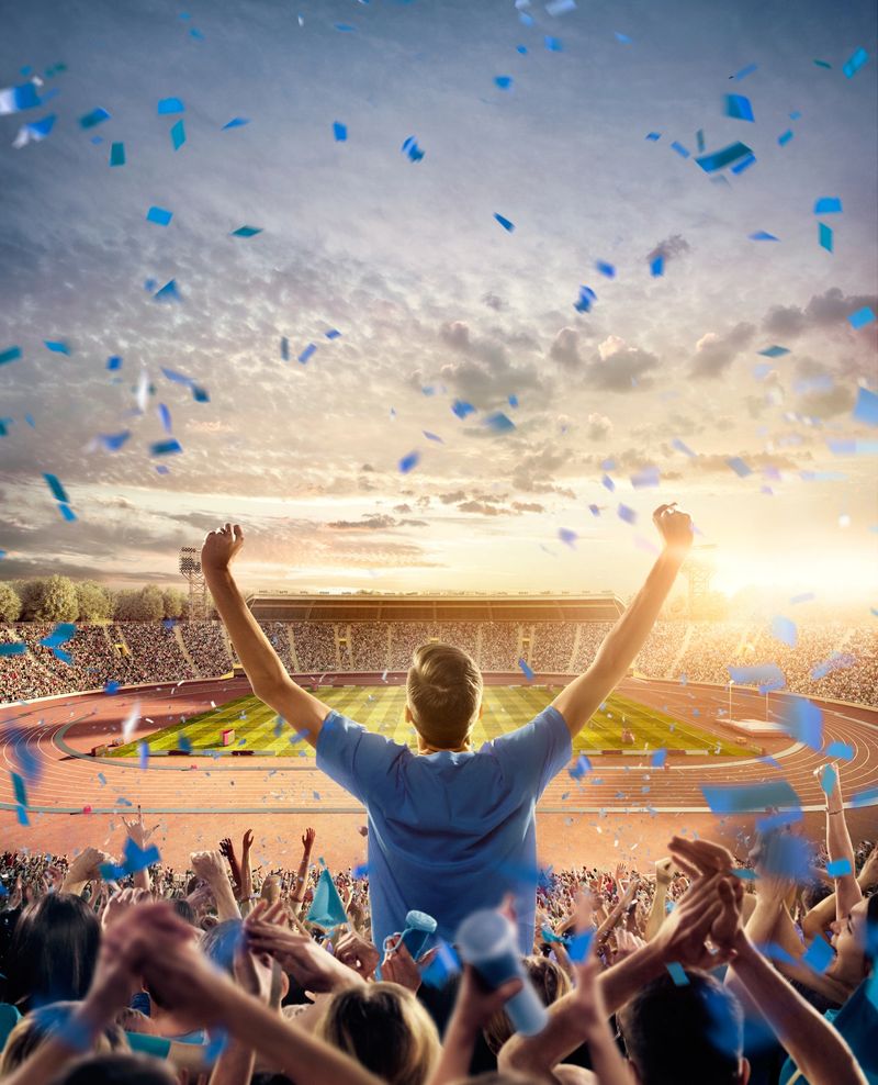 On the foreground a group of cheering fans watch a sport championship on stadium. One girl stands with his hands up to the sky. People are dressed in blue colors. A long-range shot of a stadium field, floodlights and seating. A green field, with painted white lines, is visible in the foreground. In the background are diffuse out-of-focus stadium seats. Large, bright floodlights are in the top-left and top-right corners of the stadium.
