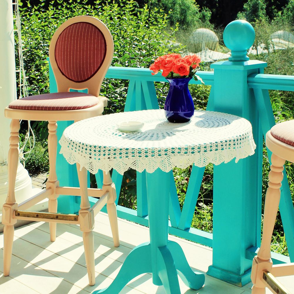 Sunny porch with turquoise railing, vintage chairs, and a lace-covered table with a blue vase of orange roses.