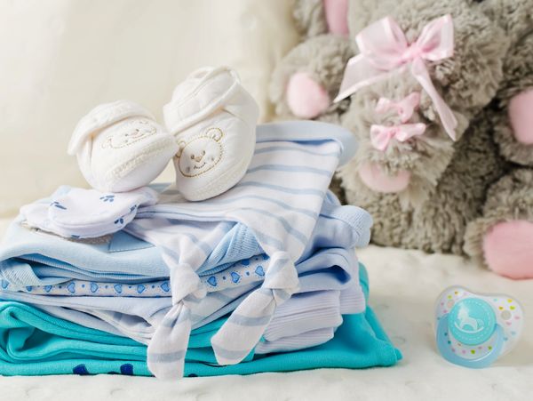 Neatly folded baby clothes with booties and a pacifier beside a plush teddy bear.