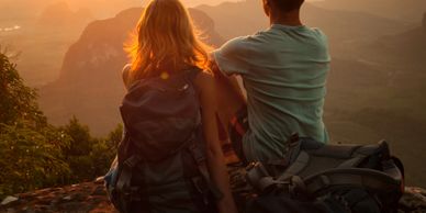 Two hikers sitting on a mountain cliff watching the sunset.