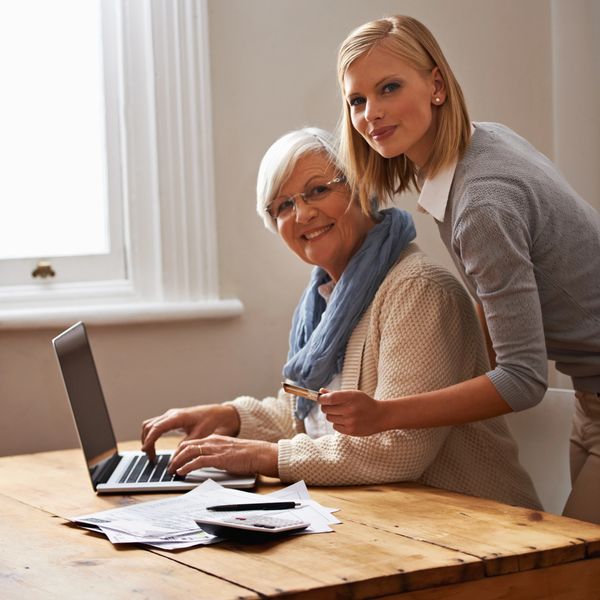 Younger woman helps elderly woman with laptop and finances at a wooden table.