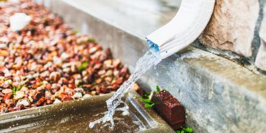 Water flowing from a downspout onto a ground surface near a rocky area.
