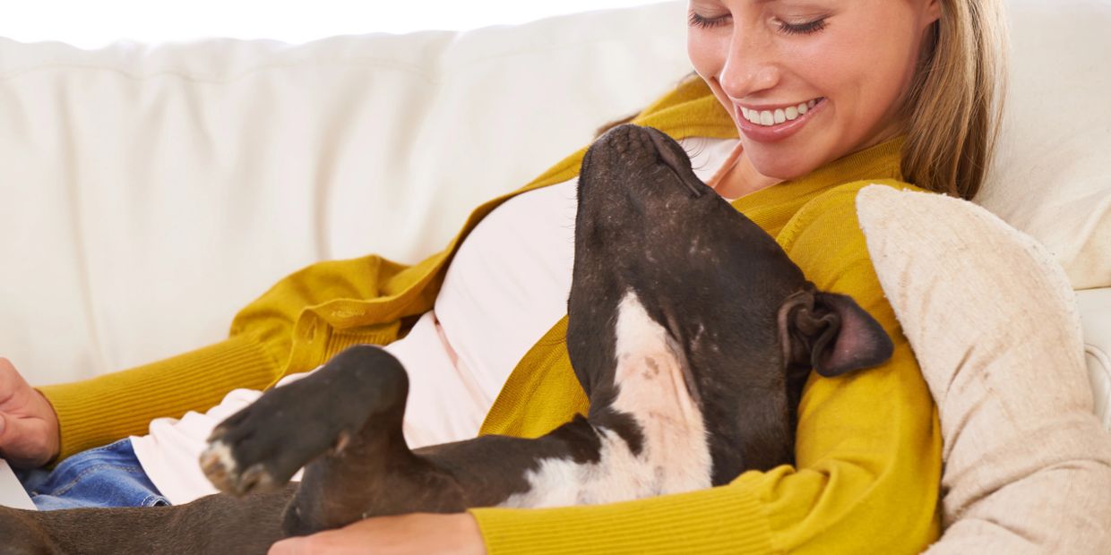 Woman in mustard sweater cuddles a black dog on a white couch.